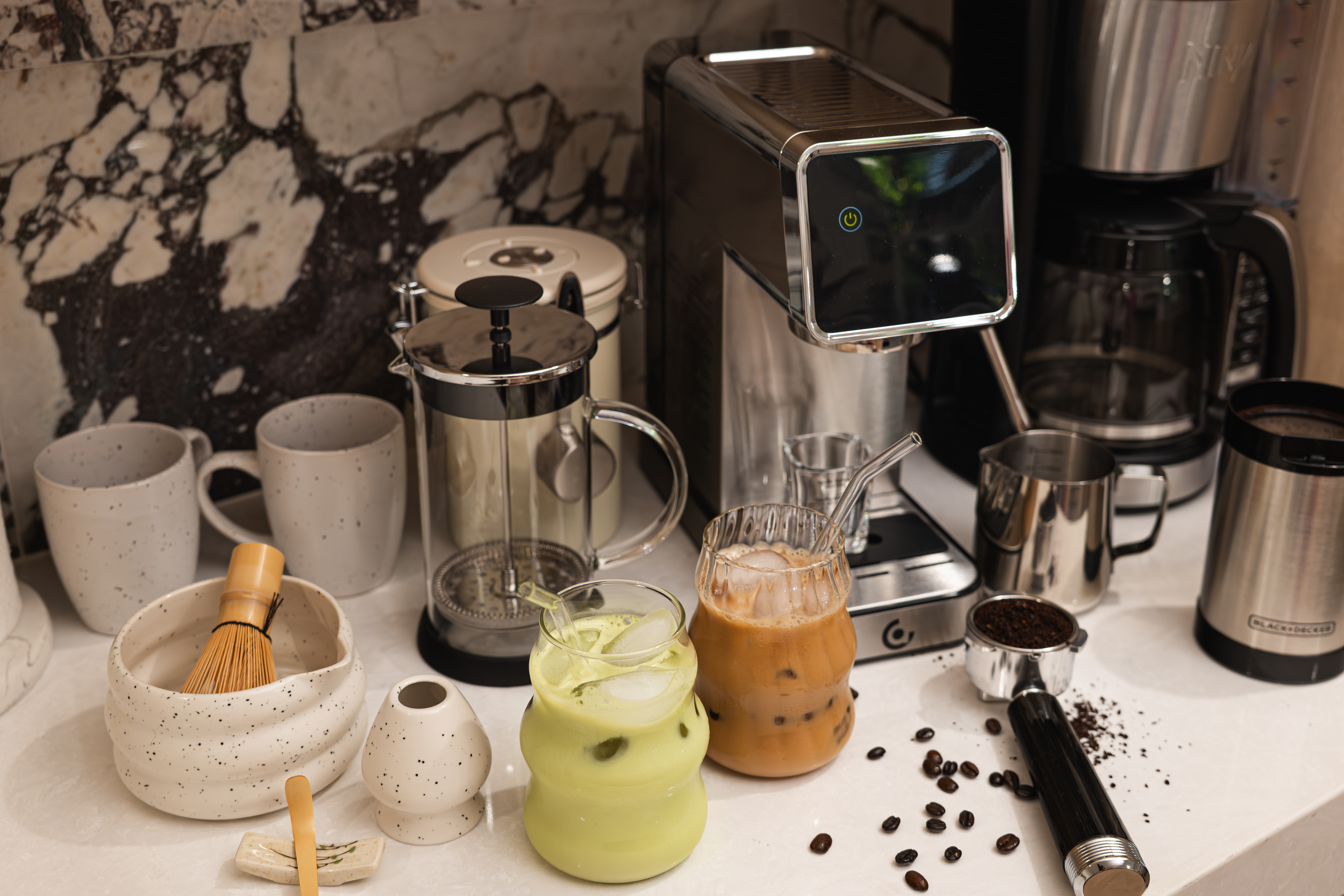Espresso machine, grinder, French press and matcha whisk set out along the marble counter